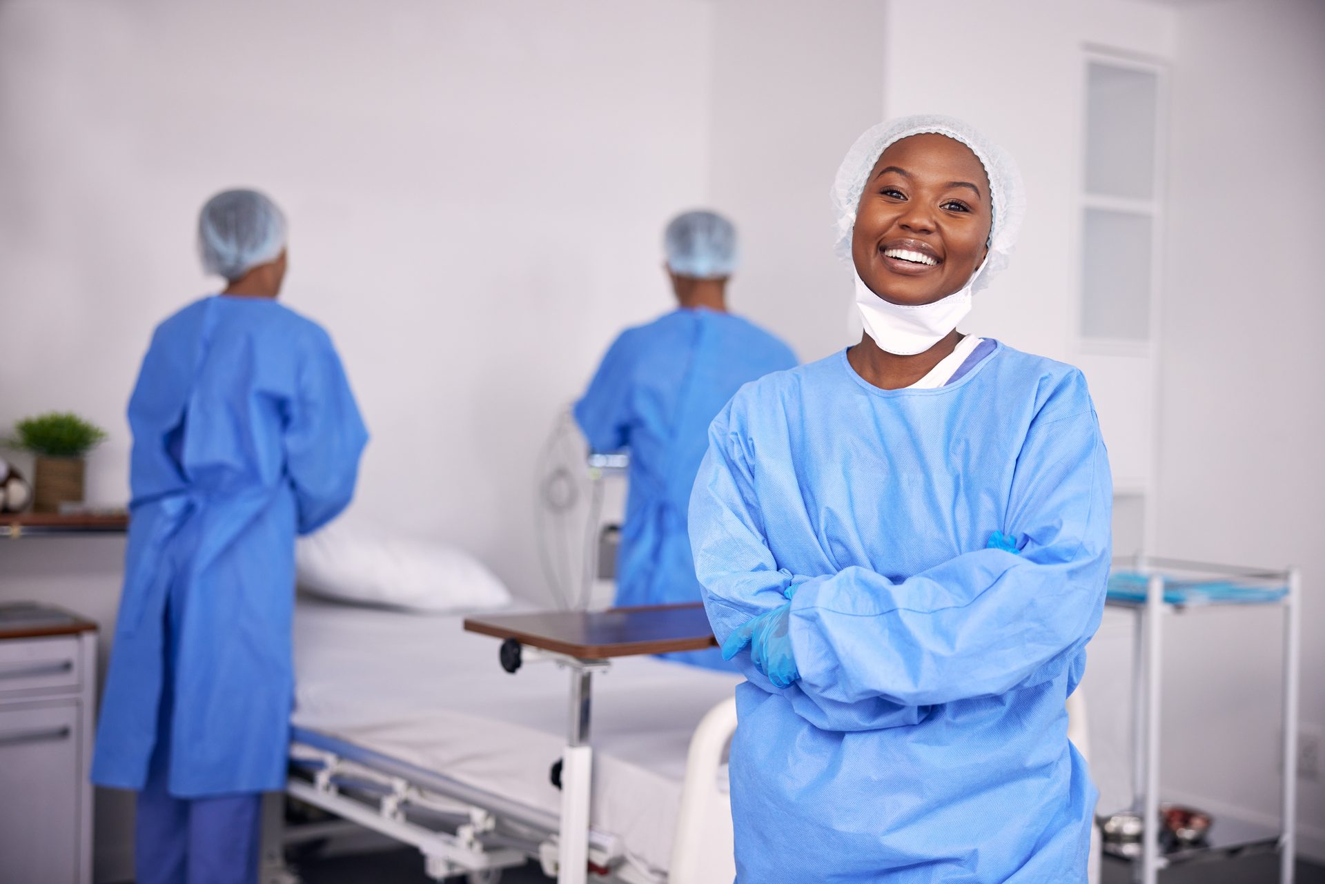 Soignante souriante en blouse, bras croisés, dans une chambre d’hôpital
